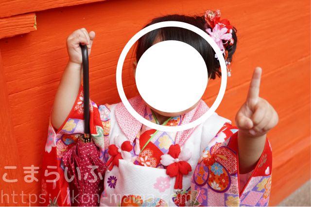 七五三で赤い壁の前に立つ着物姿の女の子が、指を立てて笑う様子。
